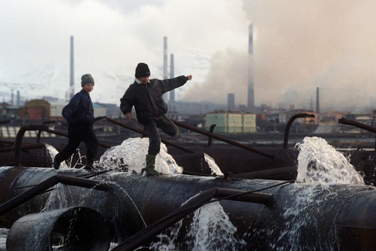 Two boys run on a pipe spurting water with smoky chimneys in the background.