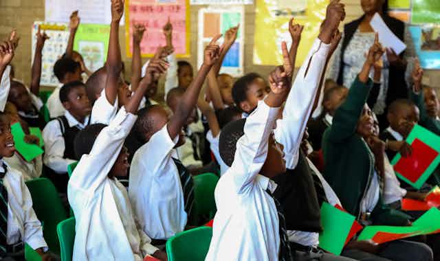 Primary school children in an overcrowded classroom with their hands up. 