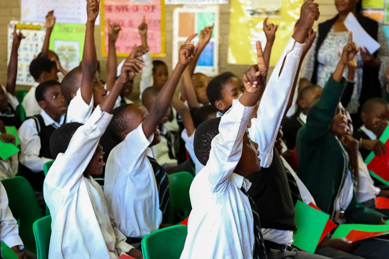 Primary school children in an overcrowded classroom with their hands up.