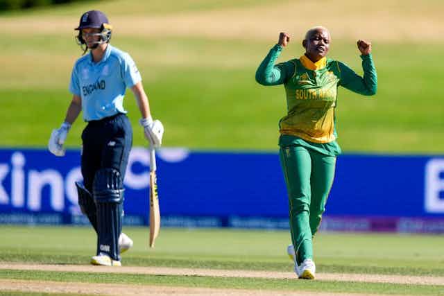 Two women in cricket suits in the colours of England (light and dark blue) and South Africa (green and gold) on a sports field. The South African holds her fists in the air, the other walks away from the wicket.