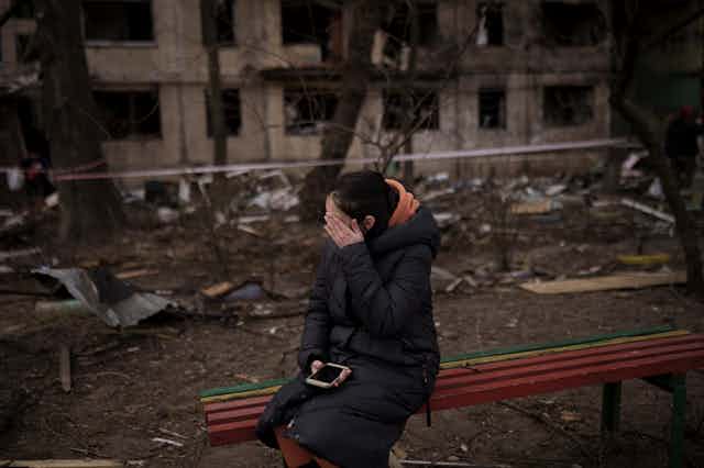 A dark-haired woman sits with her hand on her face and holding a phone on a bench outside a destroyed apartment building.