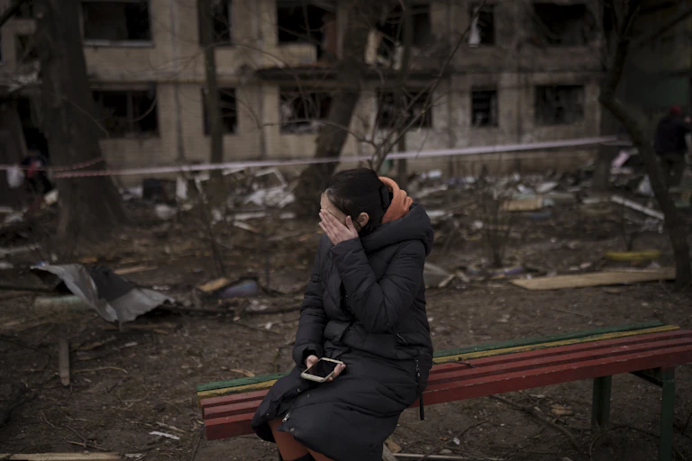 A dark-haired woman sits with her hand on her face and holding a phone on a bench outside a destroyed apartment building.