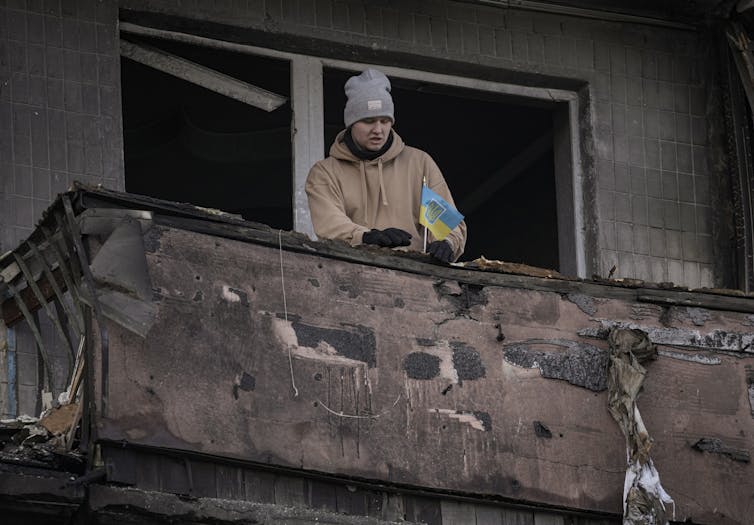 A man in a brown coat and a grey knit toque places a small Ukrainian flag on the balcony of a destroyed apartment complex.