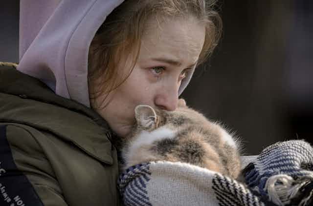 A tear streams down the cheek of a woman in a pink hoodie kissing a cat wrapped in a blanket.