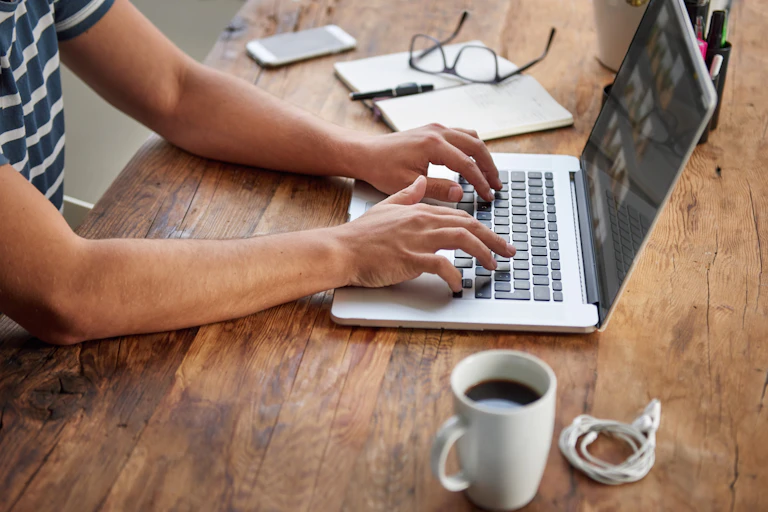 A person in a striped T-shirt typing on a laptop with a coffee and a pair of headphones in the foreground and a phone, glasses, notebook and pen in the background