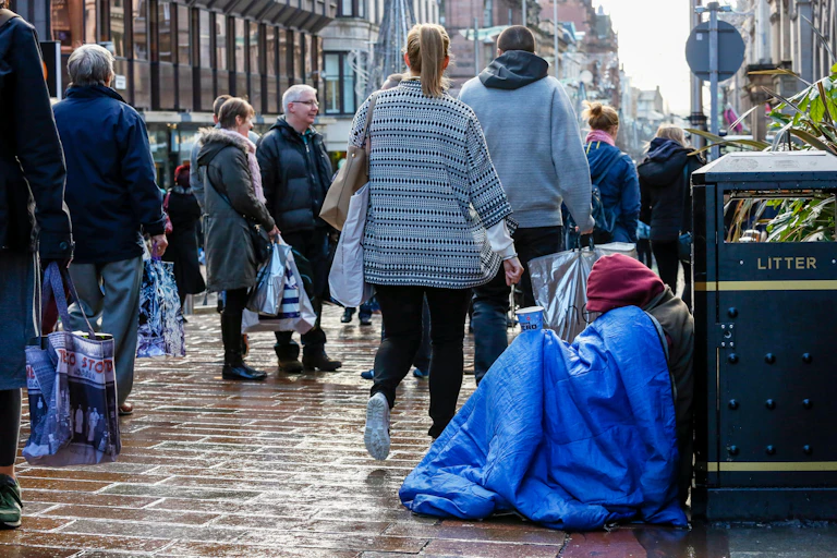A begger sits against a rubbish bin on a busy street under a blue blanket and holding a coffee cup out to passersby