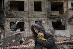 A woman in a parka carries a small orange dog in front of the rubble of a bombed building.