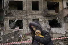 A woman in a parka carries a small orange dog in front of the rubble of a bombed building.
