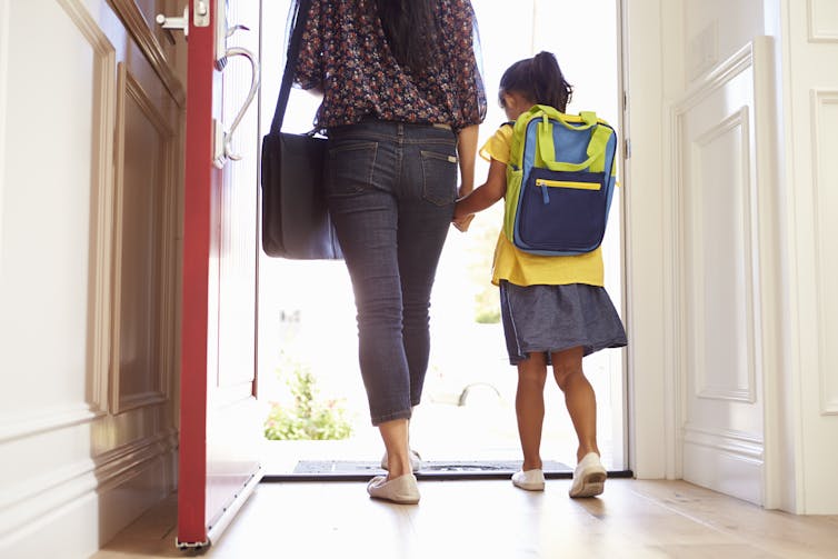 A woman with a briefcase and a child with backpack holding hands, seen from behind walking out a door.