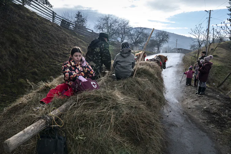 Two horses pull a cart with a doctor sitting in straw with her medical bag.