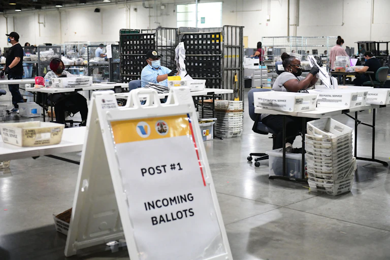 A sign says "Incoming Ballot" in an administrative looking building, with several people sitting at desks counting ballots.