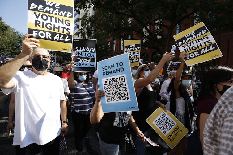People march in a street, holding up signs that say 'We demand voting rights for all' and 'Scan to demand.'