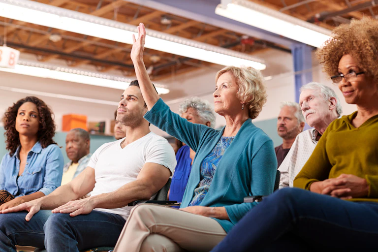 A woman, seated in chair in a small audience of other people, raises her hand