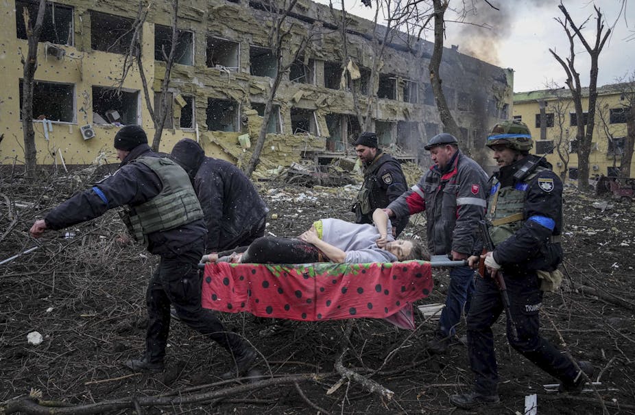 A pregnant woman on a stretcher is carried away by four men in front of bombed-out buildings.