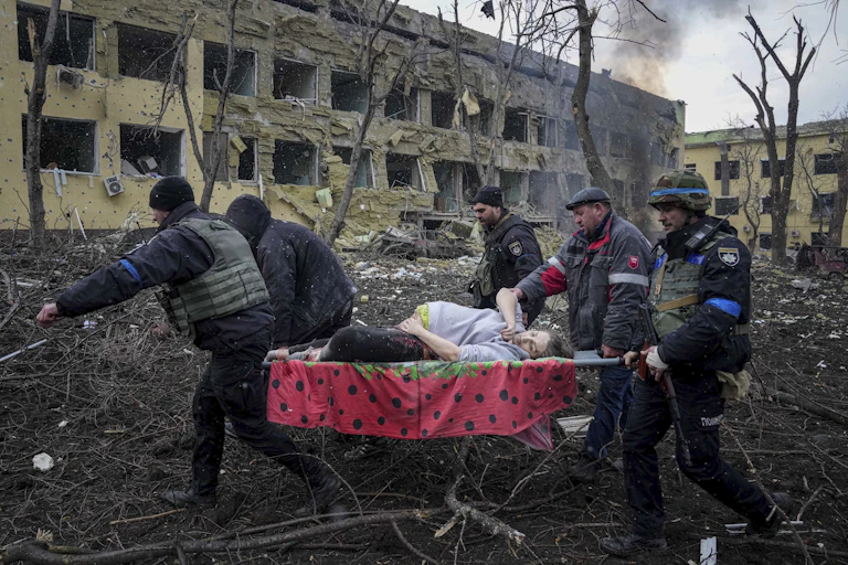 A pregnant woman on a stretcher is carried away by four men in front of bombed-out buildings.