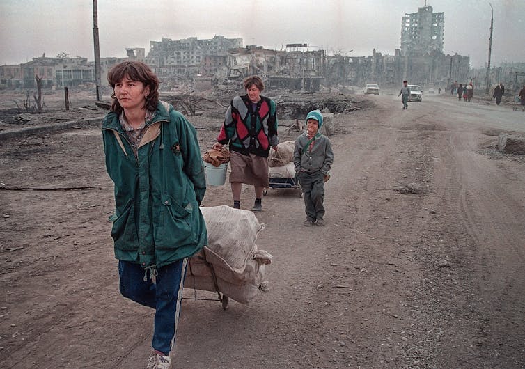 A woman dragging a cart walks away from the edifice of buildings destroyed by missiles.