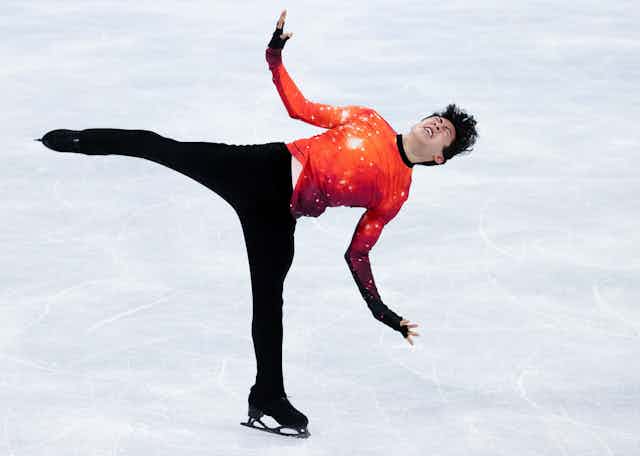 An Asian American man ice skate on one leg with outstretched arms and anguished look on his face. has his arms outstretched and 