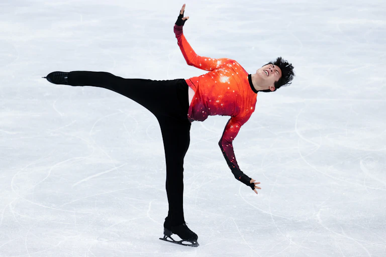 An Asian American man ice skate on one leg with outstretched arms and anguished look on his face. has his arms outstretched and