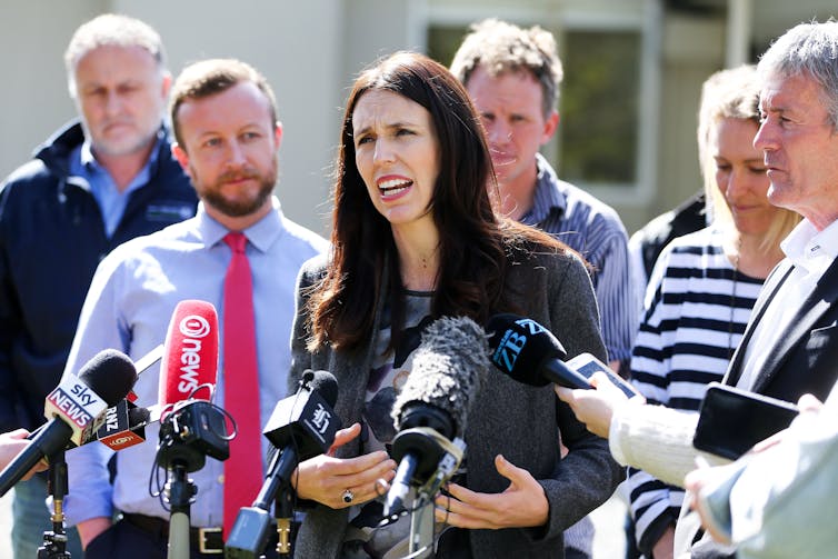 Prime Minister Jacinda Ardern speaking in front of media microphones and surrounded by other people.