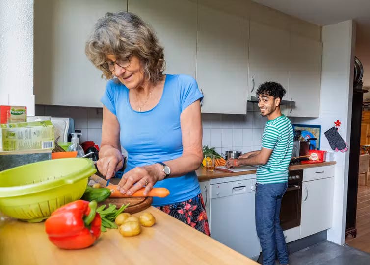 A woman chops vegetables in a kitchen with the Syrian refugee she is hosting, a young man who is also helping prepare a meal and smiling
