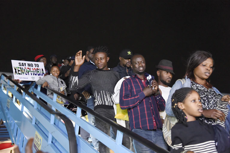 A group of men, women and children disembark an aeroplane down metal stairs, among then air hostesses holding signs that read, Say no to xenophobia'