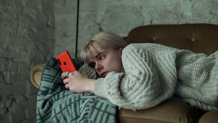 Young woman looking sad scrolls on her phone as she lays on a couch.