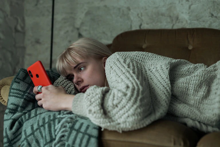 Young woman looking sad scrolls on her phone as she lays on a couch.