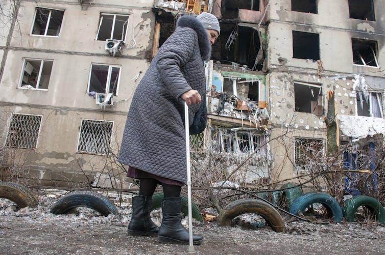 A woman with a cane walks past a destroyed apartment building.