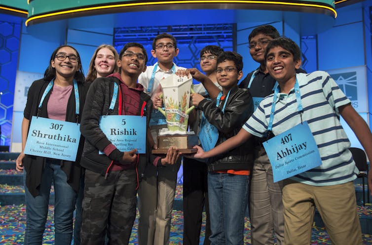 Eight people are smiling and proudly pose with the Scripps National Spelling Bee championship trophy.
