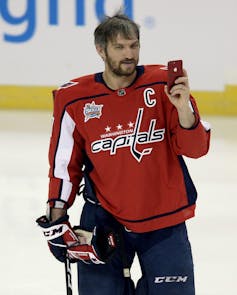 Ovechkin in a Washington Capitals jersey holds a smartphone while standing on a hockey rink.