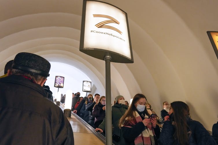 People descend on an escalator past a sign with the letter Z, a symbol of the Russian military, and a hashtag reading 'we don't abandon our own' in a subway station.