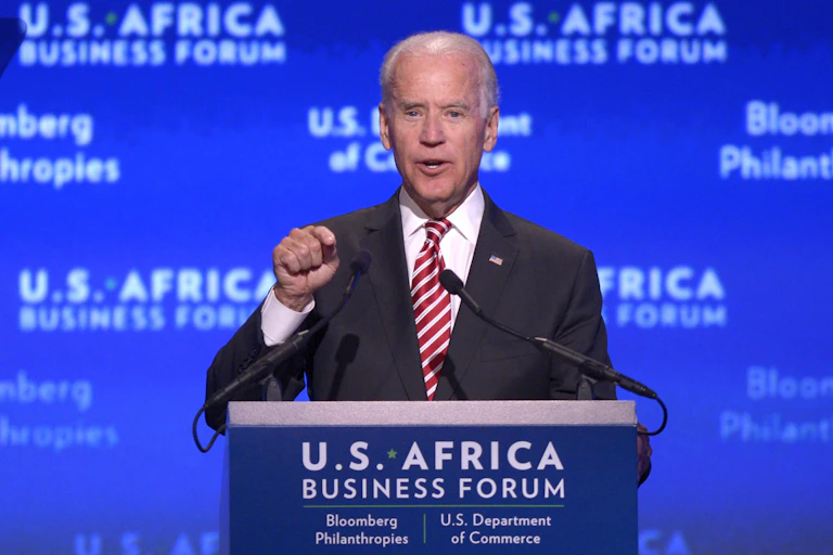 A man wearing a suit and tie points his right index finger to emphasise a point while speaking into two tiny microphones at a podium.