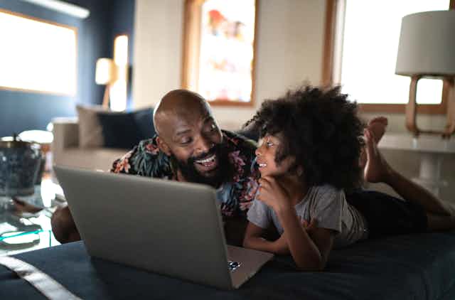 A Black man smiling with his daughter. They are both lying on their stomachs with a laptop in front of them.