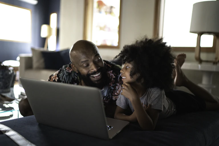 A Black man smiling with his daughter. They are both lying on their stomachs with a laptop in front of them.