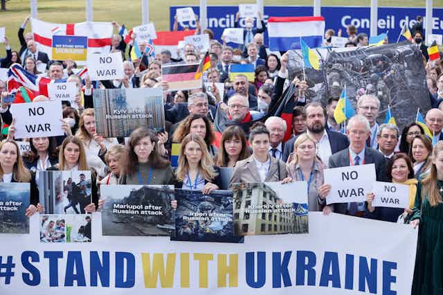 A group of people stand together behind a banner that reads 'stand with ukraine' they're holding signs that show cities after attacks and 'no war'