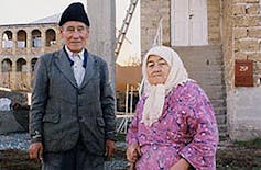 A man dressed in a suit and a black cap standing next to a woman in a white headscarf and floral pink dress.