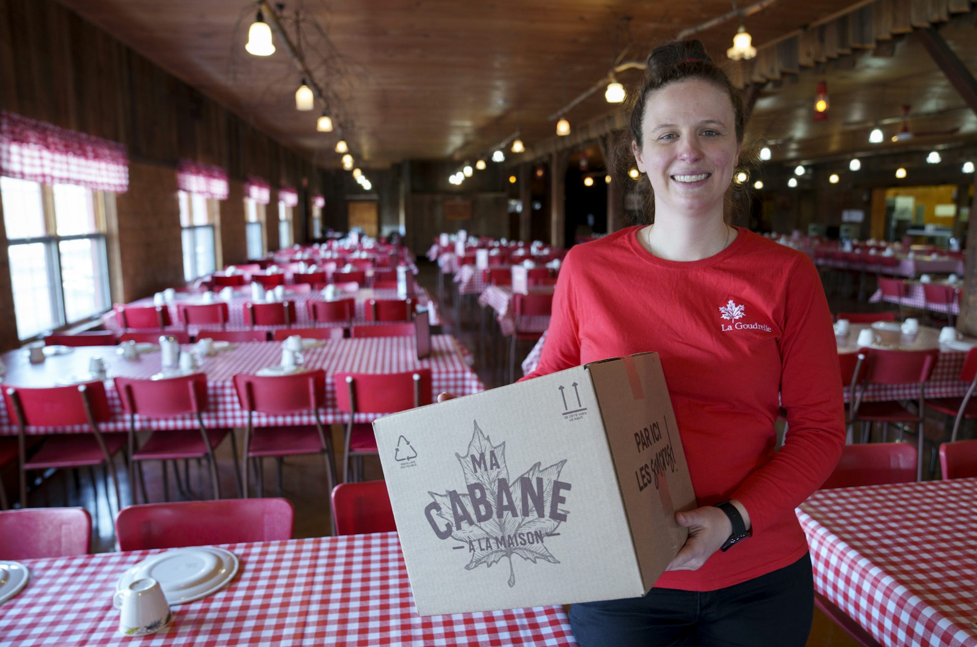 A woman stands in front of an empty restaurant. The tables are covered in red and white plaid, she's holding a box that says 'cabane' on it.