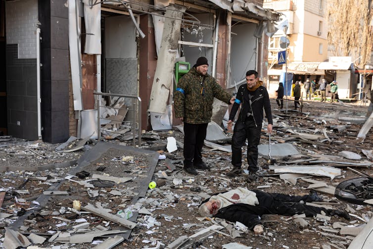 The body of a man is sprawled on the ground, with rubble surrounding him. Two men look on.