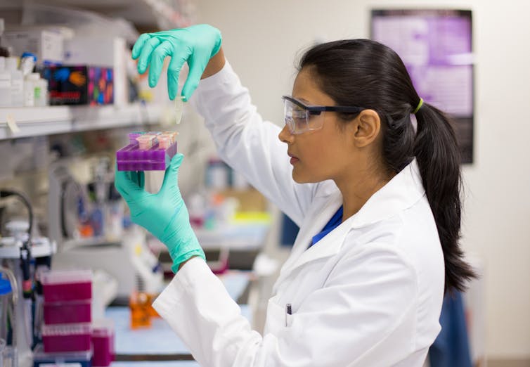 A woman wearing a lab coat, goggles and green gloves examines laboratory samples