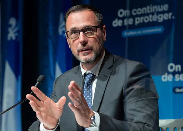 A man sits behind a mic, he's wearing a suit and brown rectangular glasses, behind him is a blue banner that reads 'on continue de se protéger' which means 'we continue to protect ourselves'