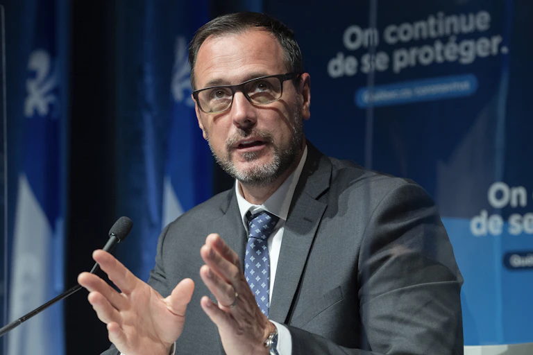 A man sits behind a mic, he's wearing a suit and brown rectangular glasses, behind him is a blue banner that reads 'on continue de se protéger' which means 'we continue to protect ourselves'