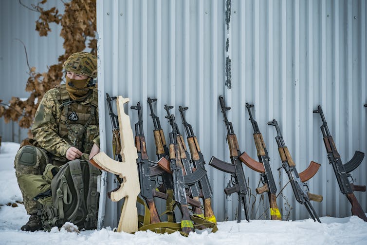 A man dressed in a camouflaged uniform inspects several rifles.
