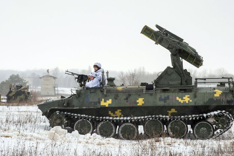 Um soldado vestido com um uniforme branco de inverno está sentado na frente de um tanque que possui um lançador de mísseis.