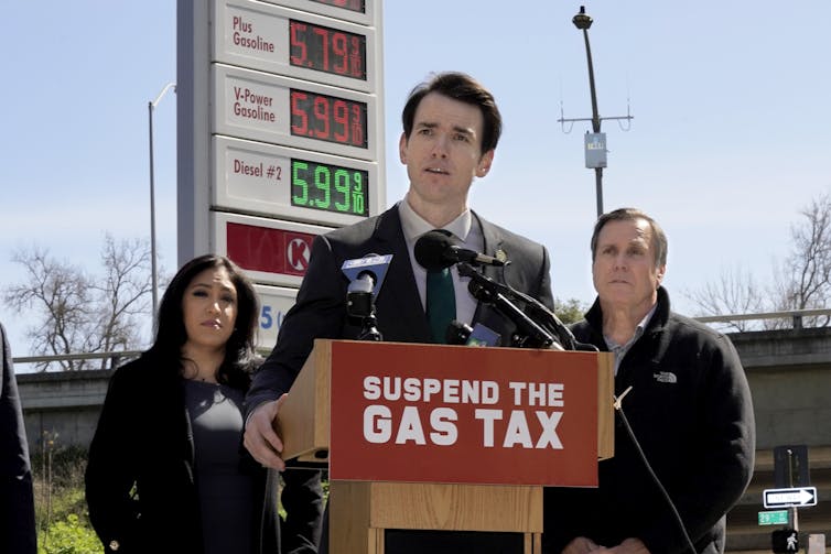 A white man wearing a suit stands in front of podium that says ‘suspend the gas tax’ as two people and a gas station sign displaying prices are in the background