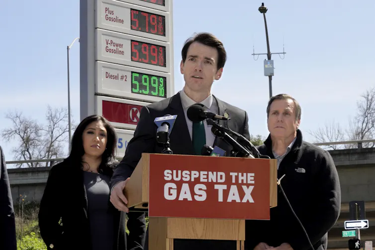 A white man wearing a suit stands in front of podium that says ‘suspend the gas tax’ as two people and a gas station sign displaying prices are in the background