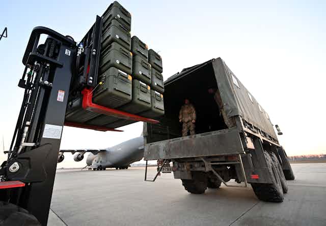A man dressed in army fatigues  stands on the back of a truck as a forklift loads boxes of weapons. 