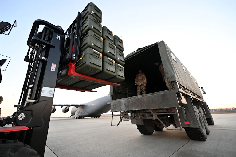 A man dressed in army fatigues stands on the back of a truck as a forklift loads boxes of weapons.