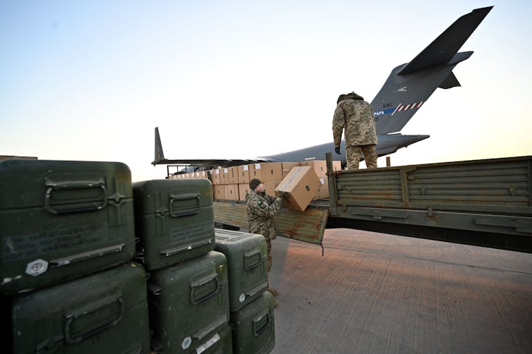 Two soldiers are unloading boxes of weapons from an airplane to a flatbed truck.