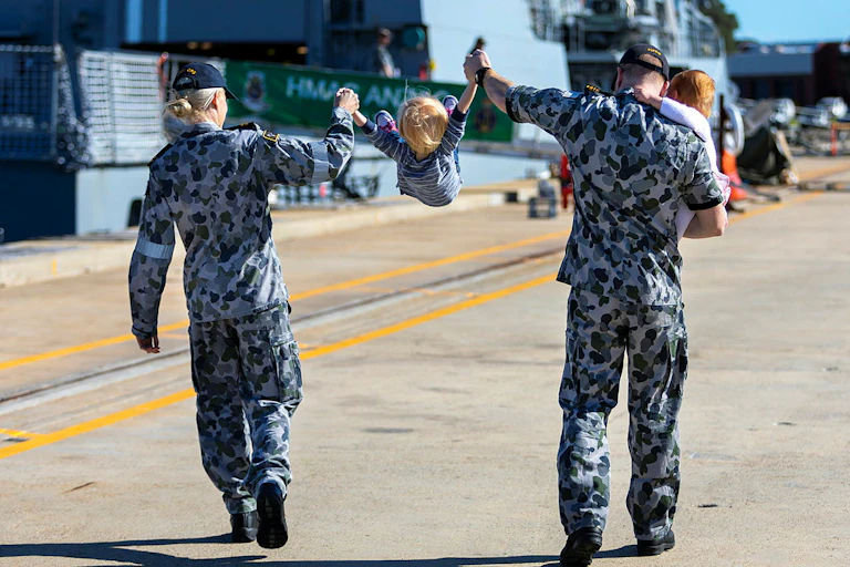 Two uniformed Navy members walking while swinging a toddler between them. An infant is being held in the arms of one member.