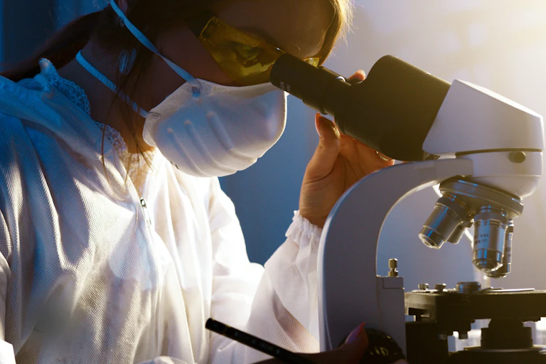 A woman wearing a face mask and protective glasses looking into a microscope